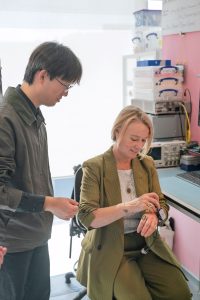 A woman sitting, tries on the D-SCAPE prototpye while a researcher watches